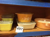 Multiple vintage Fire King and Pyrex bowls displayed on shelf with lids, colors include yellow, orange, and white.