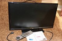 Front view of RCA 24 inch LED TV/DVD combo on carpeted floor with power cord visible. Screen black and reflective, TV logo below screen center. Remote and manual partially visible.