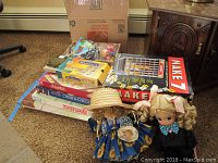 Two Precious Moments dolls with sun hats and several boxed board games stacked next to a cardboard box in a carpeted room.
