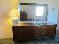 Front view of the wooden Mid-Century Modern dresser featuring six large drawers and three smaller drawers centered, with a lamp placed on top and a large mirror behind.