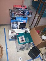 Multiple boxed items on floor including Duracraft Surround Heat heater fan model CZ2200 box, HoMedics 5 Motor Full Body Massage Mat with heat box, Lumiscope Curved Handle heat lamp box, and HoMedics Hydrogel Infrared Massager box.