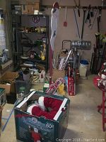 Boxed 60 inch Waving Santa figure and Christmas decor bucket in garage setting with shelves and tools in background.