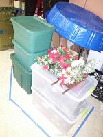 Three green and two transparent plastic storage bins stacked; artificial flowers and greenery on top; a blue plastic lid resting nearby.