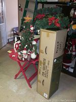 Photo of boxed artificial Christmas tree next to decorated snowman tabletop tree on red folding table