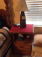 Wooden nightstand with two drawers and a red felt cover on top holding a table lamp and a clock radio, near a bed and window blinds.
