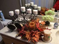 Five-armed wrought iron candelabra with white pillar candles, autumn floral centerpiece with orange candle surrounded by artificial leaves, pine cones, and berries, small vase with green hydrangea flowers, and part of two wrought iron wall candle holders visible in background