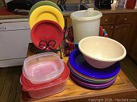 Three stoneware plates in red, yellow, and green displayed on a black wire holder stand, beige ceramic bowl, colorful plastic serving plates stacked, and large white Tupperware container with lid on counter.