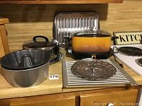 Photo showing six items on kitchen counter: two metal Bundt pans, metal cake taker, perforated steaming basket, metal roasting tray, and vintage West Bend slow cooker with glass lid and yellow-orange ceramic pot.