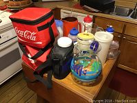 Wide shot of lot items on wooden kitchen counter showing Coca-Cola cooler, Tupperware jar, stoneware pitcher and other kitchen items