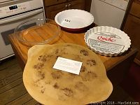 Photo showing the 15-inch pizza stone, red pie dish, white quiche dish with recipe, and Pyrex glass pie dish with lid on a wooden table.