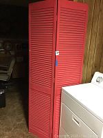 Full view of the red wooden room divider standing upright indoors by a dryer and chair, showing three slatted panels and hinges.