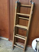 Full view of the wooden ladder folded up leaning against a wood paneled wall, showing wooden rungs and side rails with printed text.