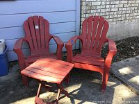 Two red plastic Adirondack chairs and a small red side table photographed outdoors next to a light blue building and brick wall.
