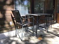 Photo showing the metal patio table with glass top positioned between two matching metal chairs outside against the house siding in sunlight and shadow.