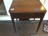 Front view of the wooden sewing machine cabinet. Medium brown finish, scratch marks visible on legs and body. Cabinet has one drawer in center and two smaller drawers on the sides.