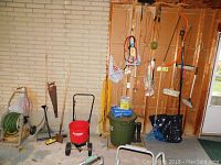 Wide-angle view of garden tools arranged against wall: hose on reel, rakes, spreader, shovel, and containers on floor