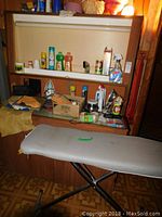 Photo showing ironing board with metal legs, two irons, various bottles of household chemicals and sprays, glue sticks, and dust rags arranged on shelves and around board.