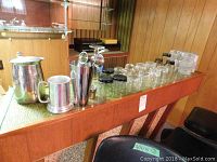 Set of glassware and barware items arranged on green surface: includes stainless steel cocktail shaker, pitcher, metal tankard, various drinking glasses, and glass jars.