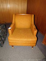 Front view of mid century modern armchair showing mustard-yellow textured fabric upholstery, wood armrests, and leg casters.