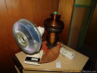 Photo showing Victorian style amber knobble glass lamp and Sears electric table fan on a table with a small wicker basket and a white corded phone