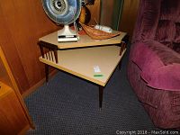 Two-tier corner table placed beside a maroon chair and against wooden paneling, showing triangular shape and melamine top shelves with wood legs.