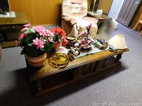 Wide shot showing all items on rectangular wooden coffee table including potted flowers, dog figurine, amber ashtray, table lamp, small decorative bowl, gift wrap, and sewing supplies.