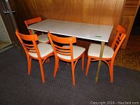 Photo showing full dining table with four orange painted wooden chairs with vinyl upholstered seats in a corner room with wood-paneled walls.