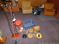 Overall view of the lot showing a black metal CD rack, orange stool, blue laundry basket, several weight plates, children's toys, and two armchairs in the background.