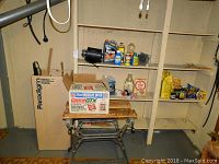 View of workbench with various cleaning product bottles and a box containing automotive oil on top of the bench. Shelves behind hold more cleaning products and miscellaneous items.