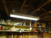 Wide view of two shelves showing assorted hardware and tools including power tools, wooden plane, tins, bottles, and painting supplies under a fluorescent ceiling light.