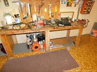 Workbench covered with assorted tools including Ashland bench grinder, hand saws, paint cans, pliers, sockets and cords. Paint and hardware underneath bench.