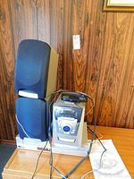 Samsung MM-29 stereo system main unit with two speakers placed on a small table with visible wood panel wall background. Unit includes connected cables and remote control on top.