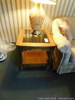 Side view of one end table showing wood frame, glass top, turned legs, and lower wooden shelf, next to a couch and a lamp on top.