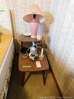 Side view of wood and Arborite end table with pink ceramic lamp, ashtrays, clock radio, and other small items placed on table tiers.