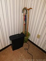 Paper shredder placed on carpeted floor next to a green bucket holding three walking canes against wallpapered wall