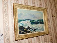 Shot of the full framed seascape painting showing ocean waves and rocks with cloudy sky, hanging on wall with patterned wallpaper.