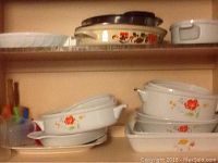 Two shelves of ceramic and metal cookware. Top shelf has stacked beige metal cooking pans with lids featuring floral band decoration. Bottom shelf shows white ceramic cookware and baking dishes with red and orange floral prints, including casseroles and rectangular pans.
