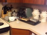 Photo showing corner countertop with covered canisters, metal bread box, cookware, glassware, and ceramic dishes