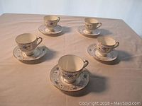 Seven demitasse cup and saucer sets arranged on a table showing blue floral pattern with gold trim.