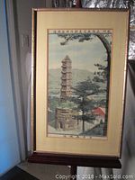 Full view of framed silk tapestry showing tall tower over a village and valley, framed in a wooden frame with beige and glass cover.
