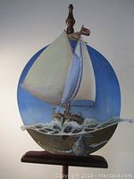Full view of circular wood oil painting depicting a sailboat sailing on waves with a minke whale underwater, blue sky background, attached wooden finial and base.