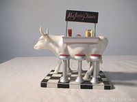Front view of the MOO Jersey Diner Cow Parade resin figurine showing the cow, diner stools, countertop with diner items, and the sign on top.