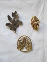 Three vintage pins on white background: a gold-tone MFA marked pin with ornate interlocking rings, a leaf shaped pin, and a round pin depicting nativity scene.