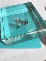 Two small sterling silver open heart earrings with a curl detail shown on a clear acrylic block with Tiffany & Co. logo.
