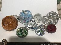 Full view of all ten paperweights arranged on white background showing variety of colors, sizes, and designs including large bubble-like clear glass paperweight, deep red sphere, blue and pink speckled piece, and others.