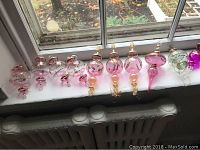 Wide view of 12 vintage style glass Christmas ornaments in pink and purple hues with gold accents placed on a window sill.