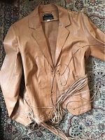 Front view of tan leather jacket laid flat showing fringe belt and floral cut-out details on cuffs and lower front.