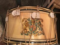 Close-up of drum body showing detailed hand-painted British royal crest featuring lion and unicorn, leather tabs holding rope tensioning, and black stripe accents on cream-colored wood body.