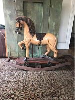 Front and side angle view of wooden rocking horse with carved brown mane, saddle, and mounted on rocker base with wheels. Missing tail visible.