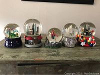 Front view of all five musical snow globes lined up on a distressed wooden surface, showing diverse themes including Macy's parade, Harry Potter, and decorative deers.